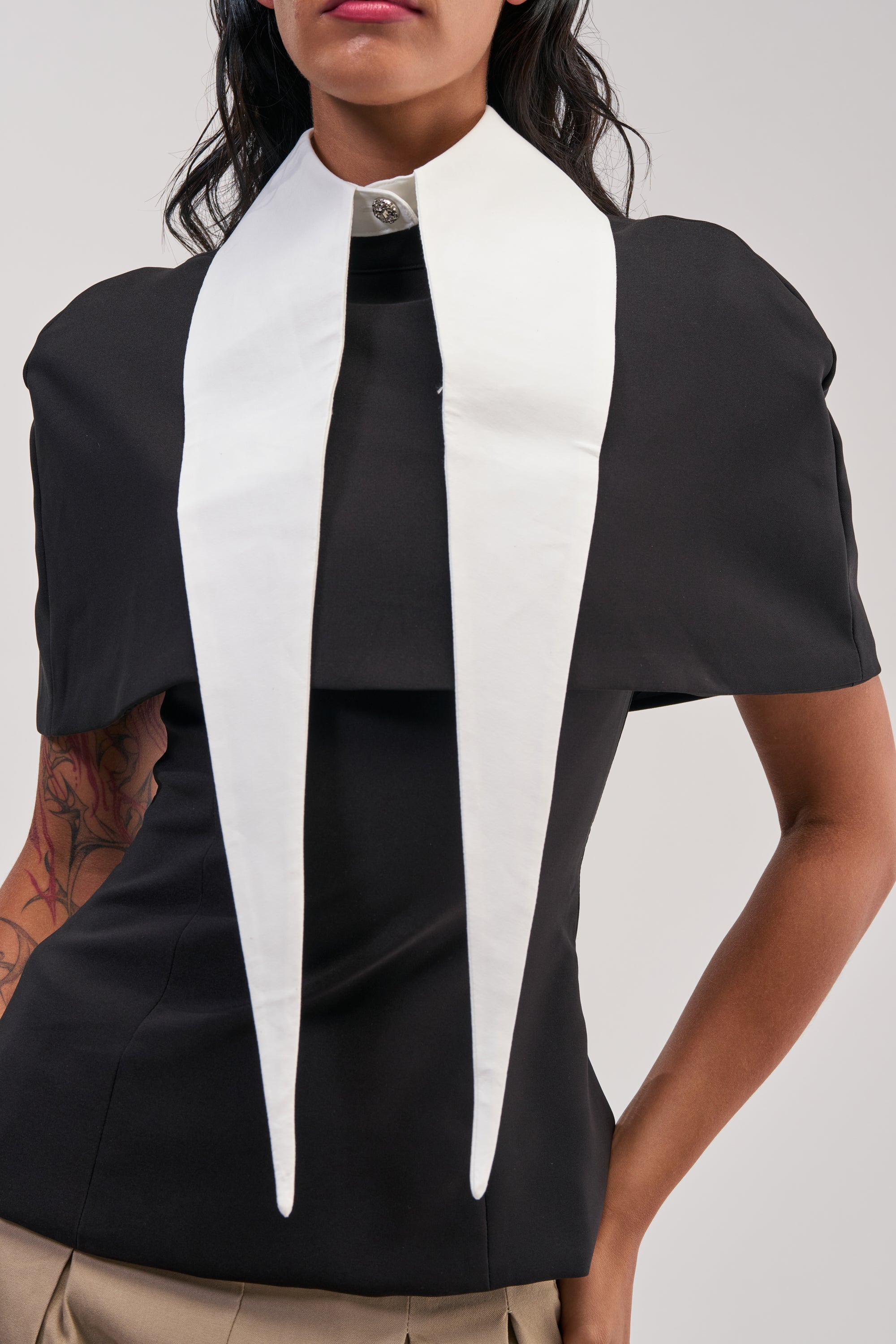 Wearing the DOUBLE DAGGER COLLAR—a long, pointed white collar with two prominent, structured cotton ends—over a black short-sleeve top and beige pants, a person showcases office siren style. One arm is bent at the waist, highlighting a visible tattoo on their left arm.