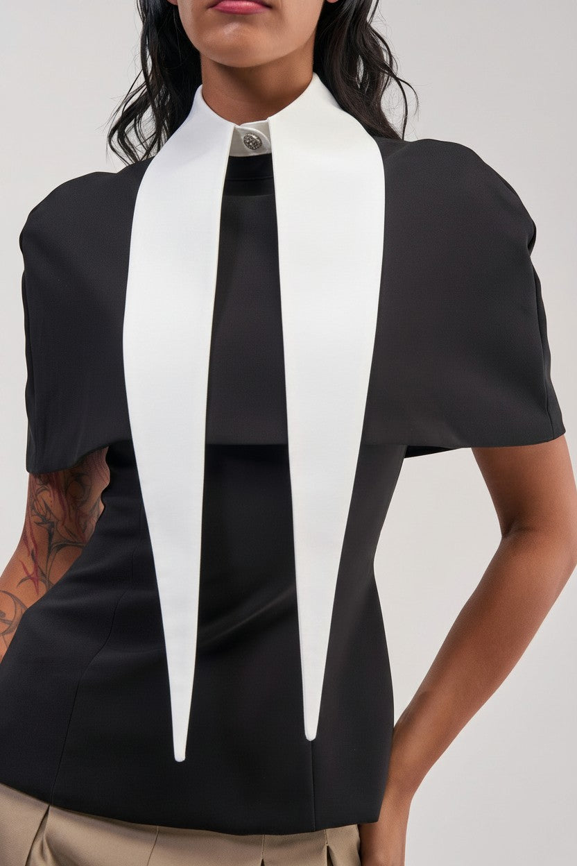 A person is wearing the DOUBLE DAGGER COLLAR, a black top distinguished by its structured cotton collar with two long, pointed white panels draping down the front. Their bent arm reveals a tattoo, while the plain neutral background adds to the look’s understated yet polished office siren vibe.