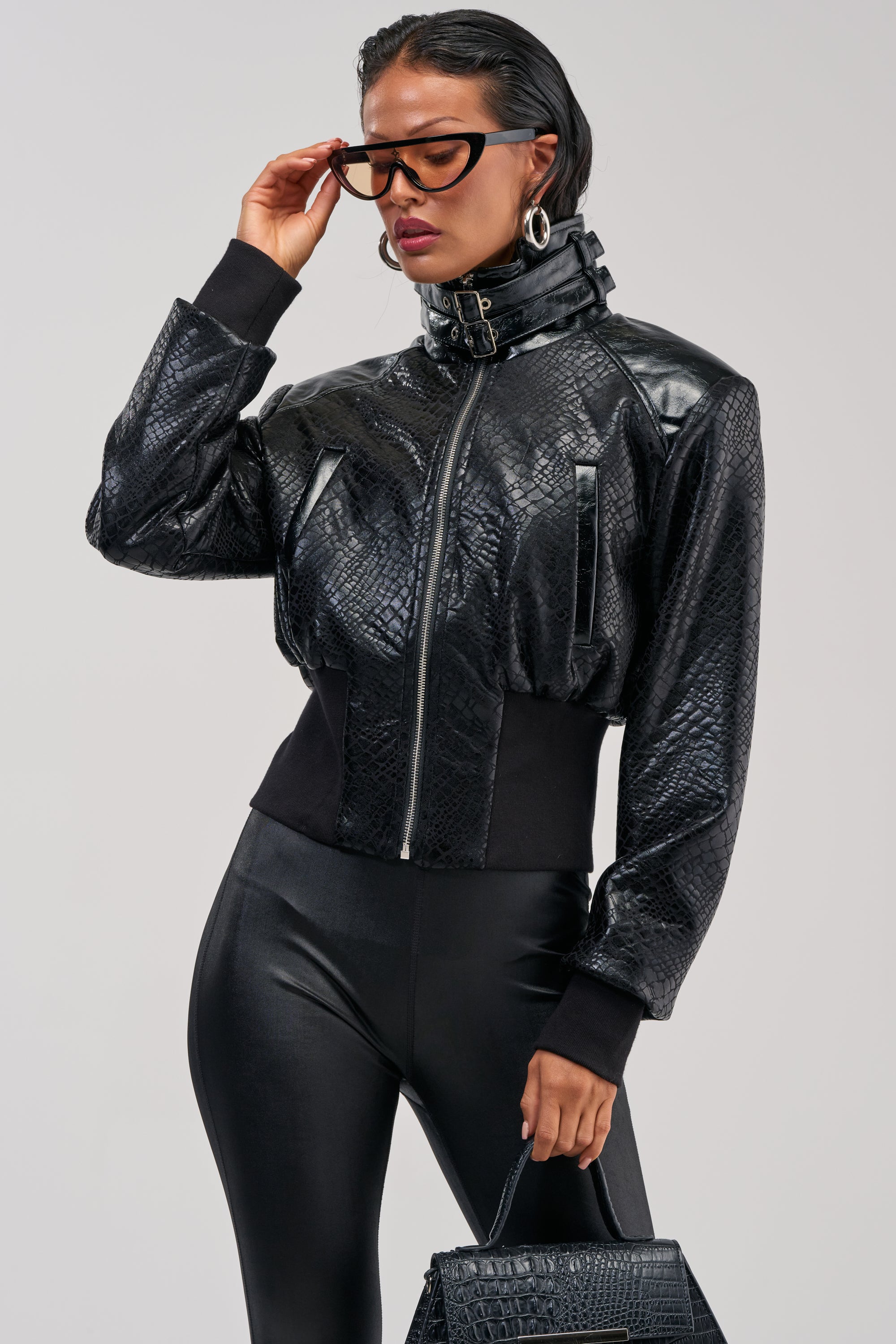 Wearing the NAOMI FUNNEL NECK BOMBER—a black jacket with a croc-embossed texture—a person pairs it with black leggings, large hoop earrings, glasses, and carries a matching textured handbag against a plain background.