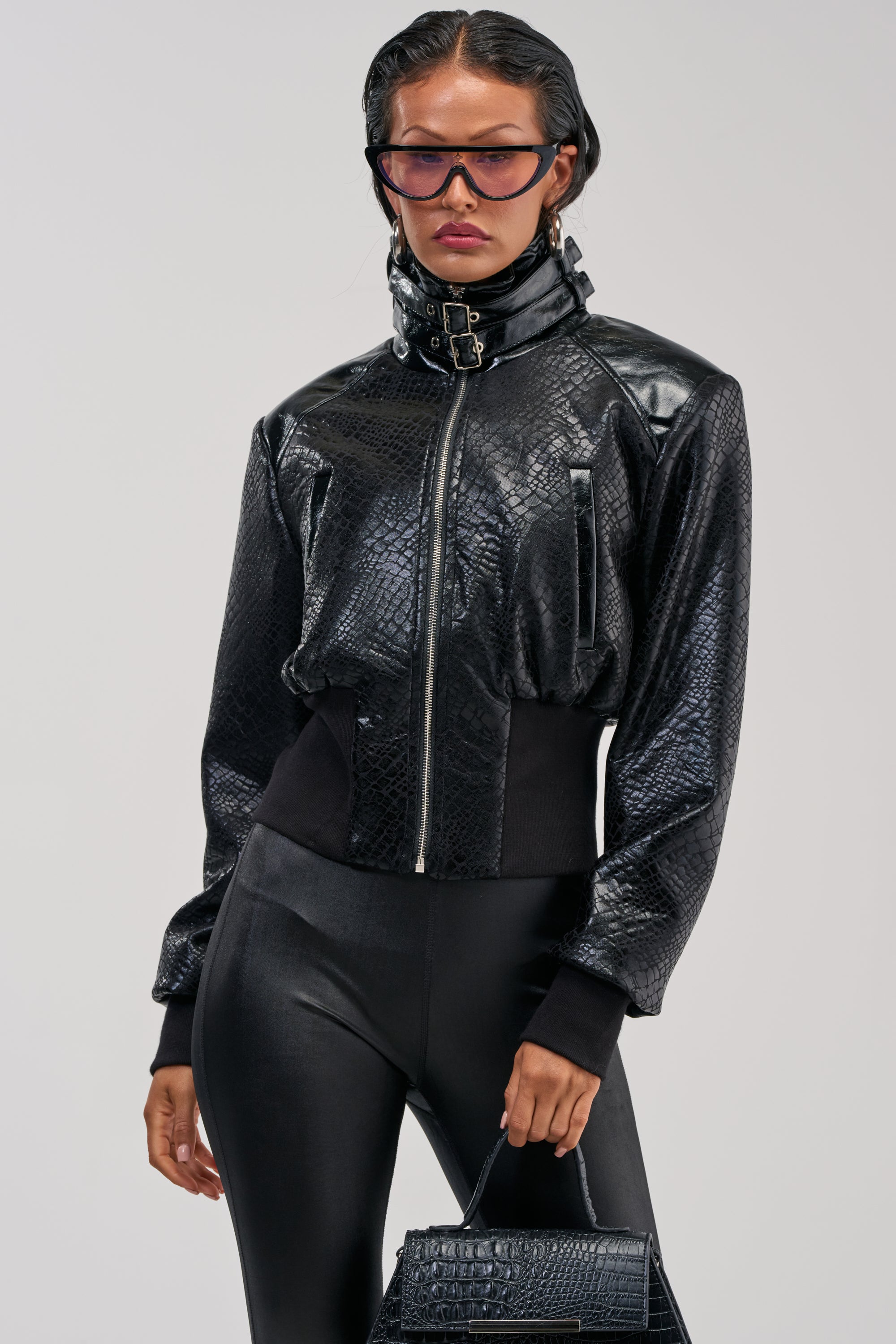 A woman stands against a plain background wearing the NAOMI FUNNEL NECK BOMBER, a black faux leather bomber jacket featuring a croc-embossed texture and a high buckled collar. She pairs it with black leggings, oversized sunglasses, and carries a black handbag.