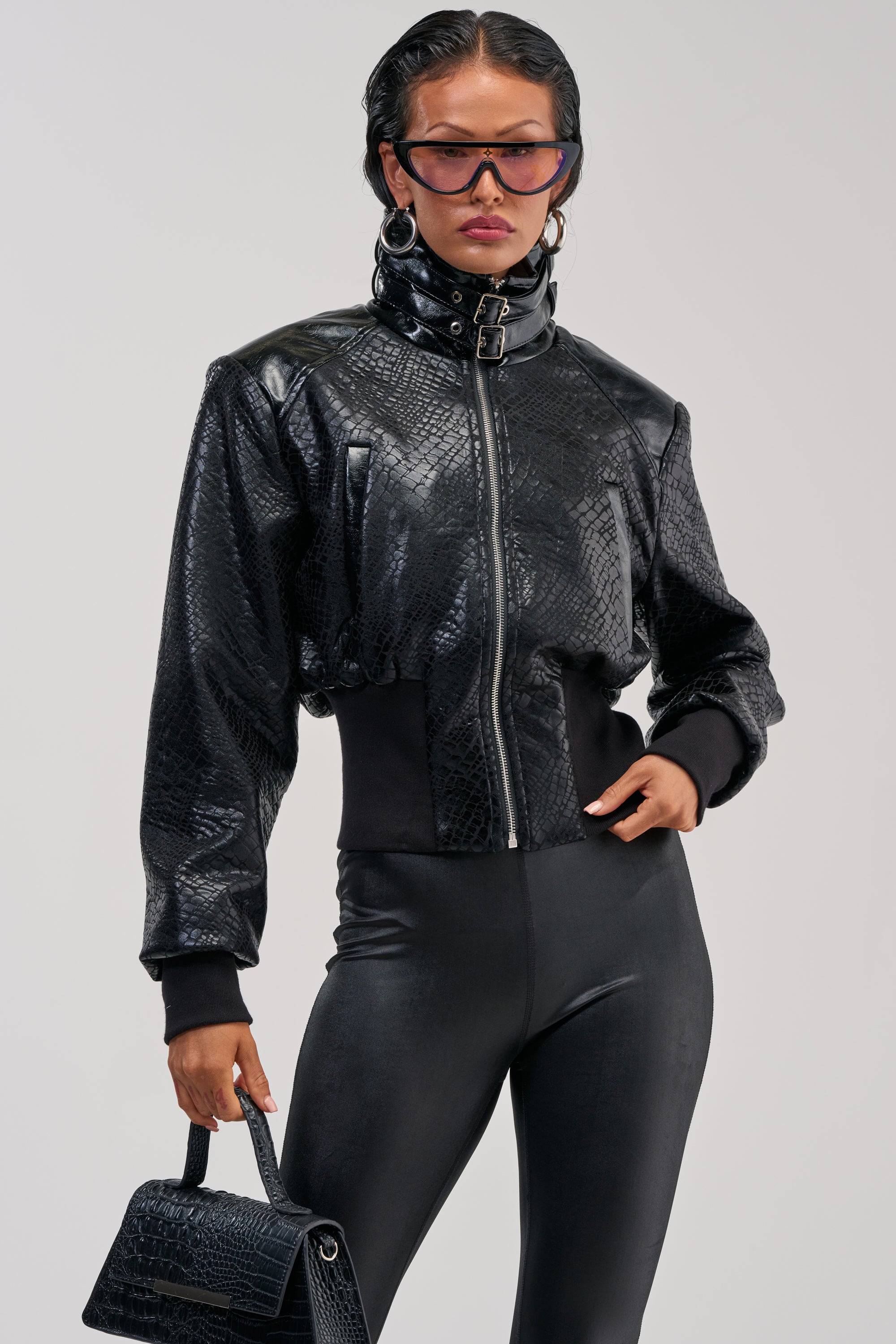 A woman wears the NAOMI FUNNEL NECK BOMBER—a shiny black faux leather bomber with a cropped silhouette—paired with matching pants and a croc-embossed textured handbag. She accessorizes with sunglasses and hoop earrings, her hair slicked back, as she poses confidently against a plain background.