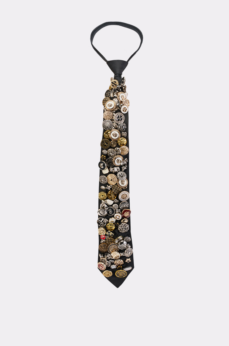 The PUSHING MY BUTTONS TIE is a black satin tie densely adorned with an eclectic mix of colorful and mixed metal buttons in various shapes and designs, displayed against a plain white background.