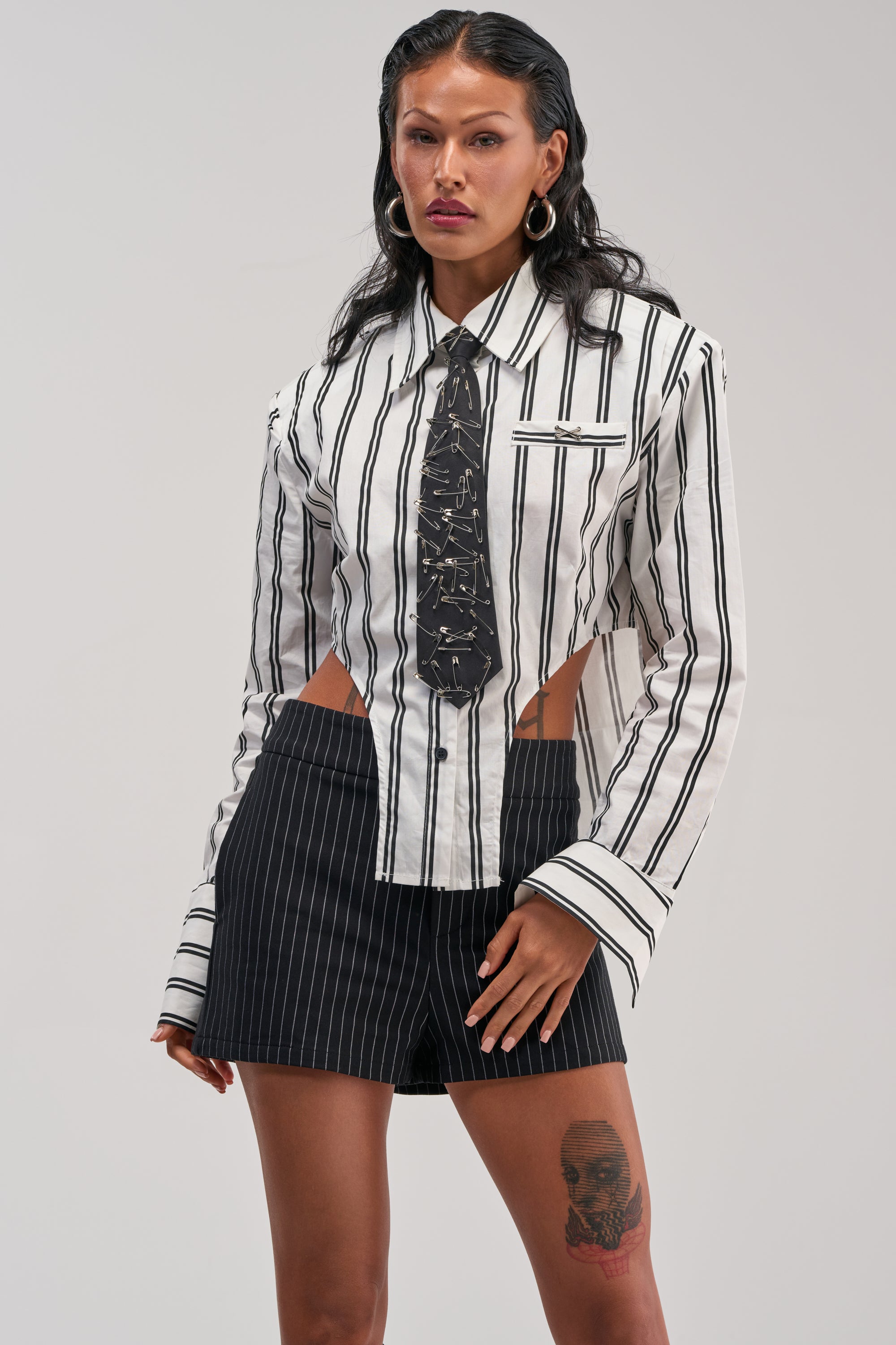 A woman with long dark hair and a visible thigh tattoo poses against a plain background, wearing the ON TOP POPLIN BUTTON DOWN—a black-and-white striped poplin top featuring cut-out sides—styled with a patterned black tie and black pinstripe shorts.