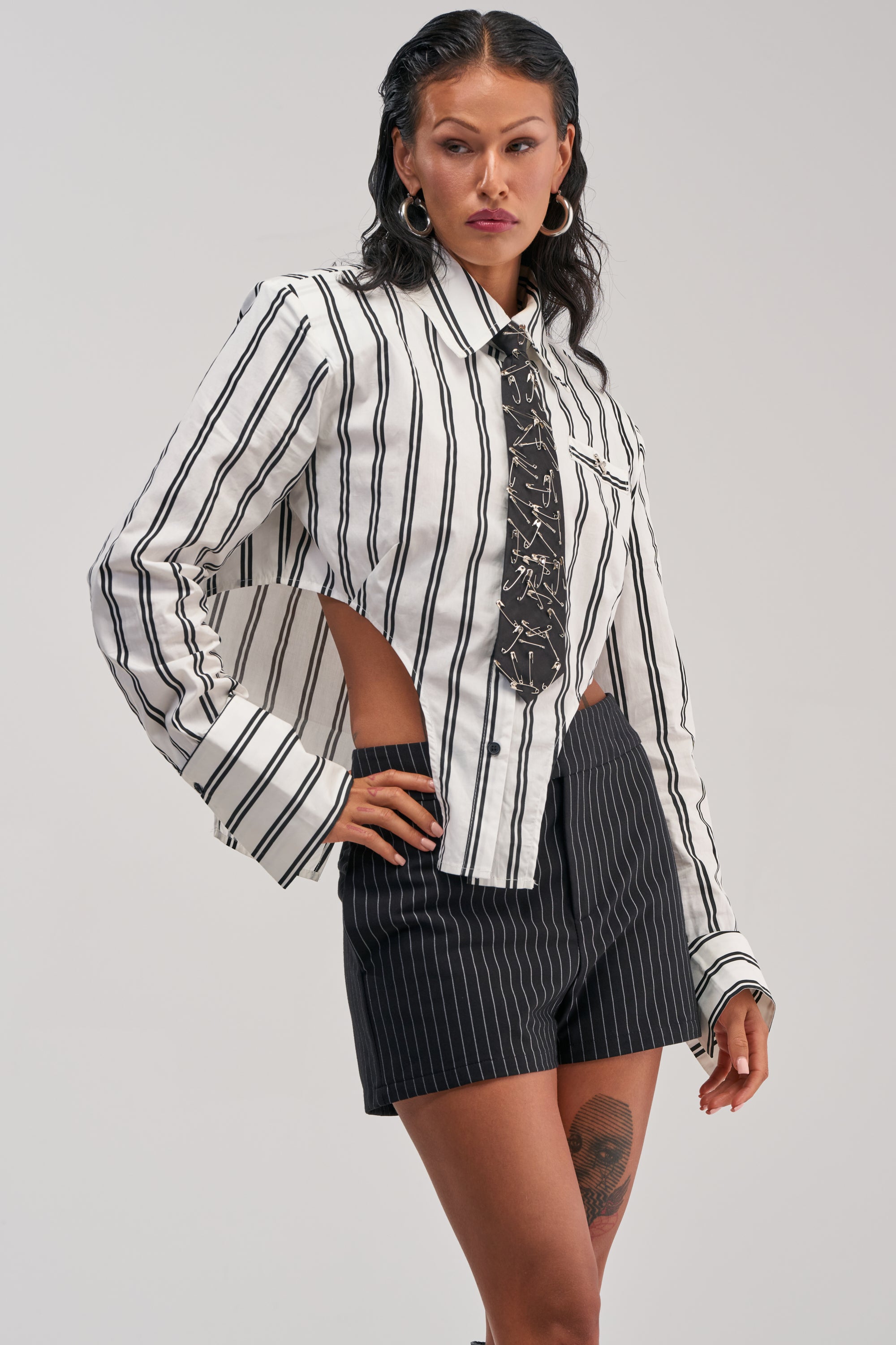 A woman poses in the ON TOP POPLIN BUTTON DOWN, a white and black striped top featuring cut-out sides and a patterned tie, paired with black pinstripe shorts. She accessorizes with hoop earrings and displays a thigh tattoo.
