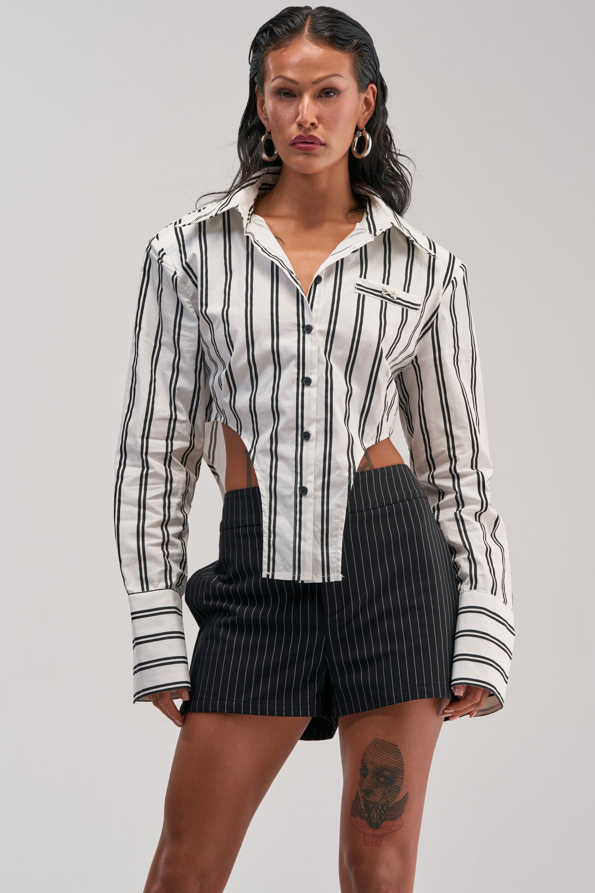 A woman is wearing the ON TOP POPLIN BUTTON DOWN, a black and white striped poplin top featuring cutouts at the waist and long cuffs, styled with high-waisted black pinstripe shorts. She has long dark hair, hoop earrings, and a thigh tattoo.