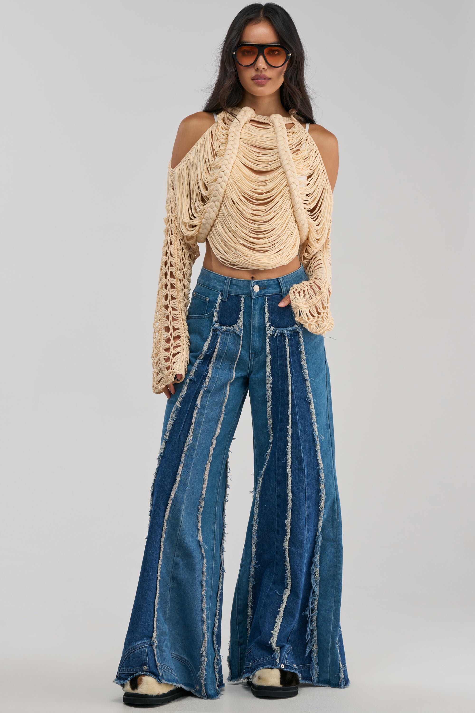 Wearing the “Denim For Days Distressed Denim” high-waisted flare jeans with exposed seams and frayed details, a woman stands against a plain background, styled with large sunglasses and a beige crochet top featuring cut-out shoulders.