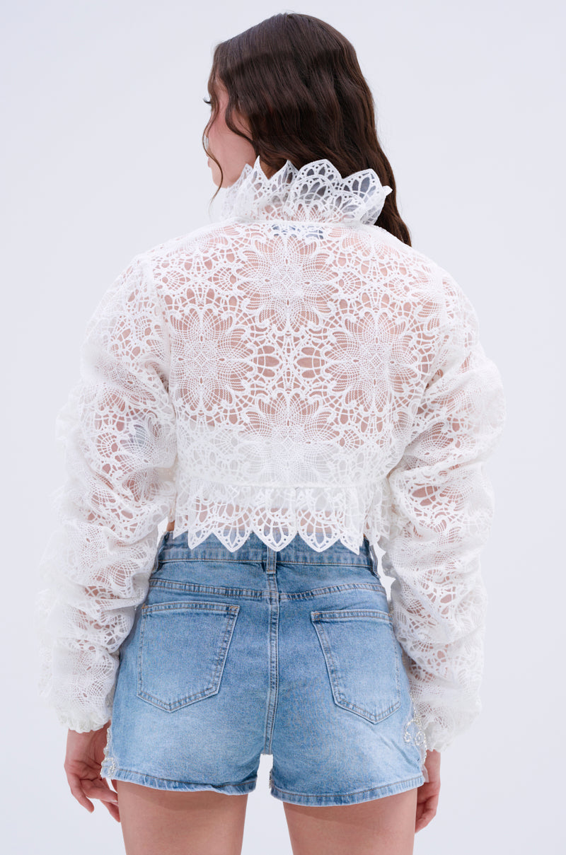 A person with long brown hair is shown from the back, wearing the SWEET THING LACE SHRUG—a sheer blouse with a ruffled collar and ruched sleeves—paired with high-waisted blue denim shorts against a plain background.