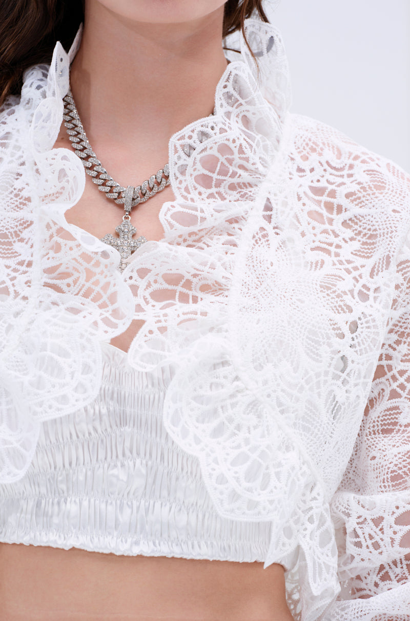 A person wears the SWEET THING LACE SHRUG with ruffled edges over a white ruched bandeau, accessorized with layered silver chains and a cross necklace. The cropped silhouette contrasts against the plain, light background.