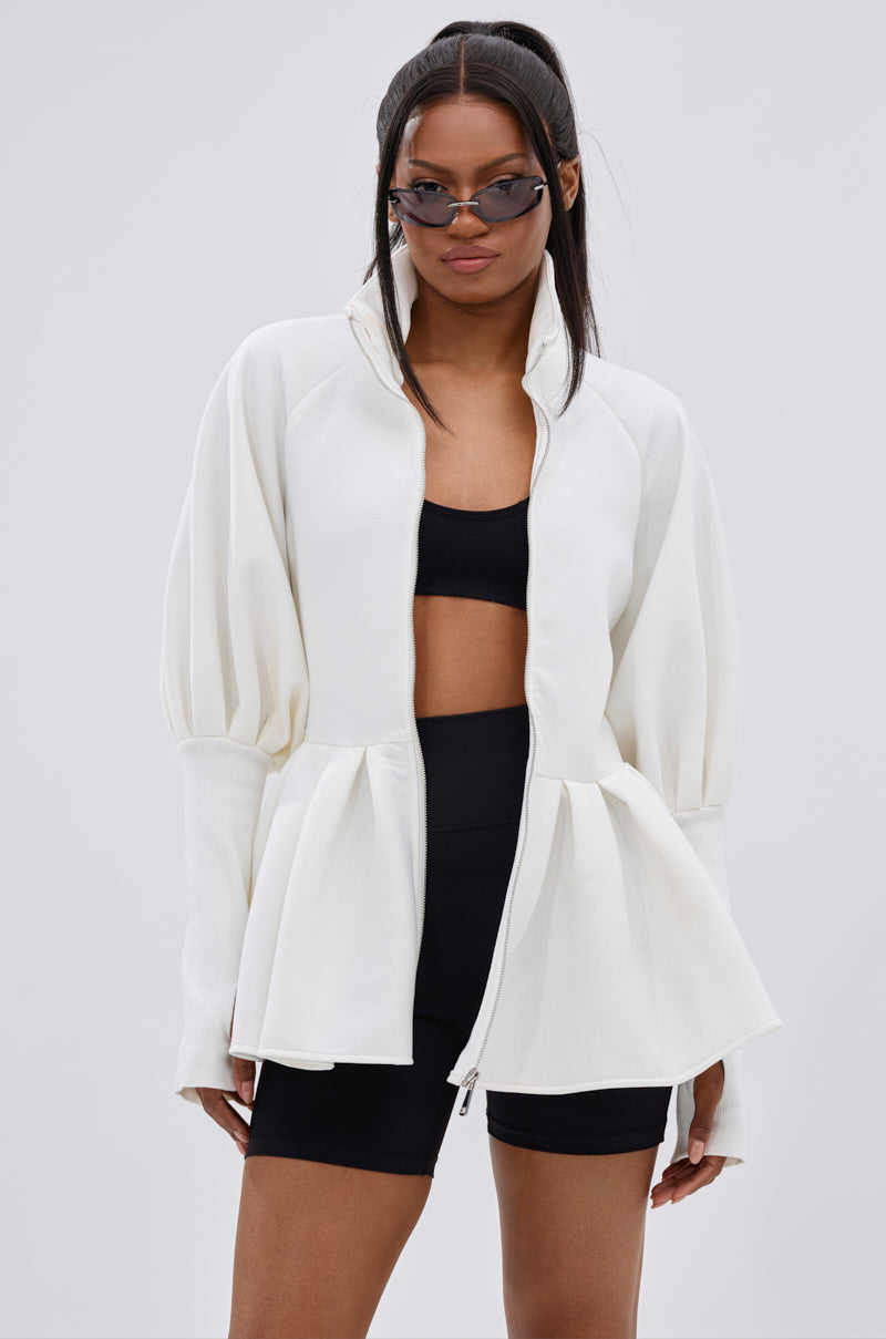 A woman showcases effortless street style in the TWIN FLAME PEPLUM BOMBER IN WHITE, paired with sunglasses, a black crop top, and black athletic shorts against a plain light background.