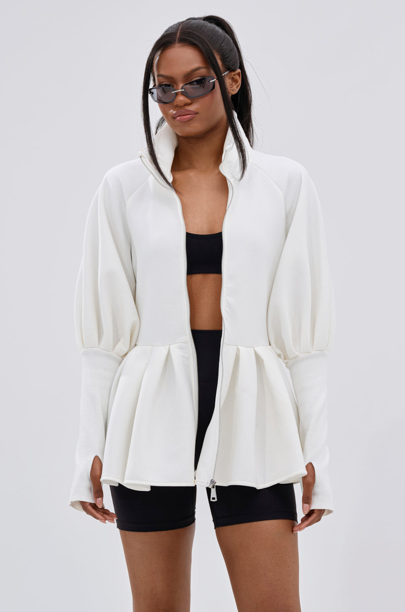 A woman in black sunglasses, a crop top, and shorts completes her look with the TWIN FLAME PEPLUM BOMBER IN WHITE, exuding effortless street style as she poses against a plain light background.