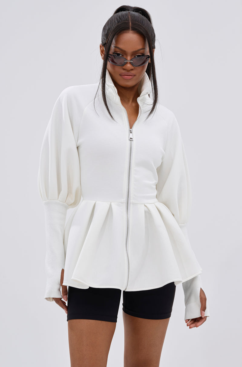 A woman wears cat-eye sunglasses, black fitted shorts, and the TWIN FLAME PEPLUM BOMBER IN WHITE with voluminous sleeves, standing against a plain light gray background.