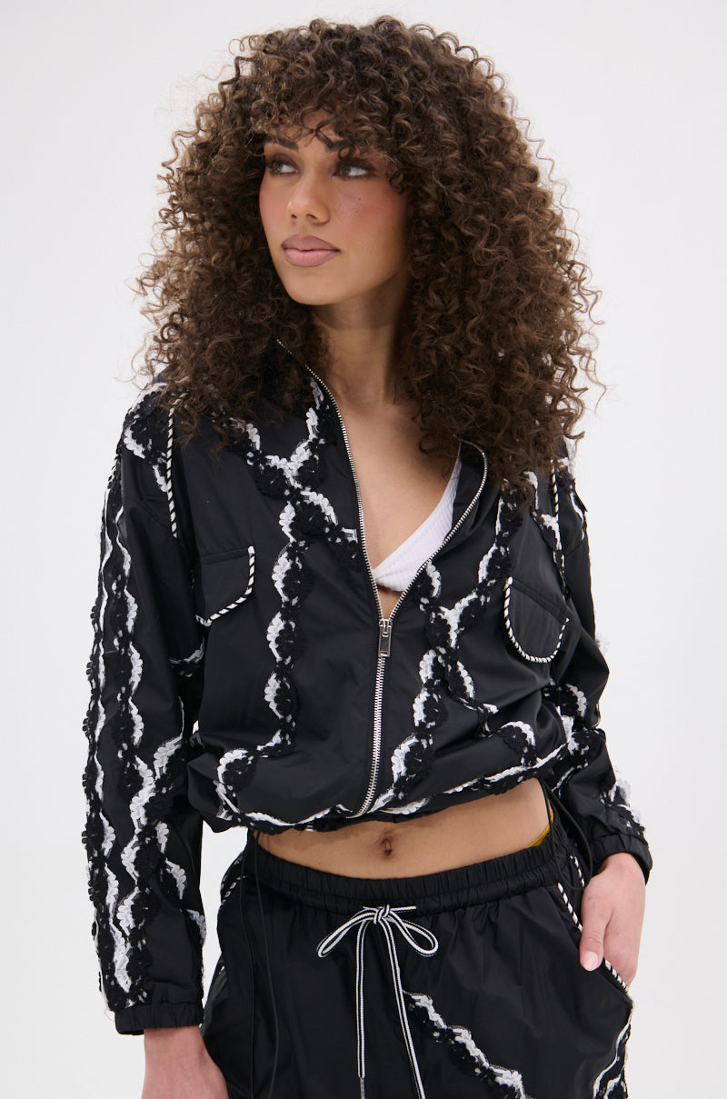 A woman with curly hair models the STRIKE OUT LACE TRIM BOMBER in a black and white pattern with matching drawstring pants over a white top, gazing to the side against a plain background.