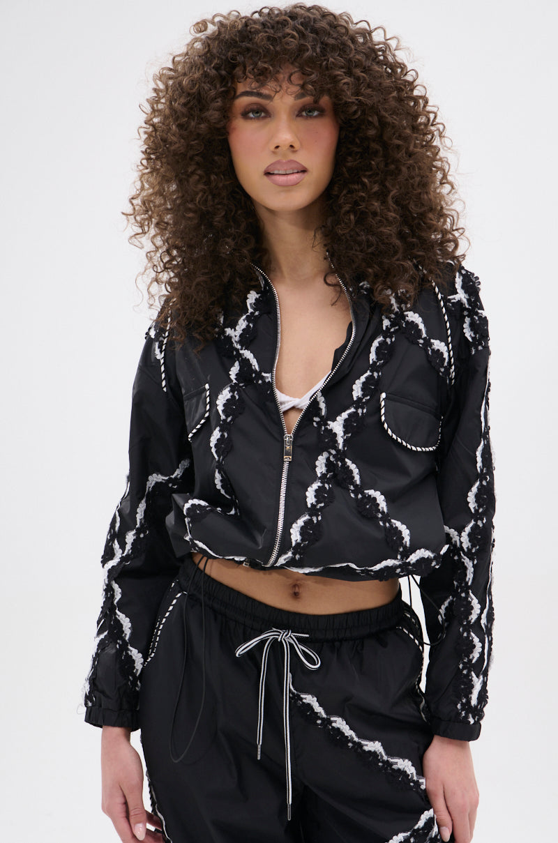 A woman with curly brown hair models the STRIKE OUT LACE TRIM BOMBER and matching tie-dye pants with white drawstrings, showcasing a street-casual look against a simple light backdrop.