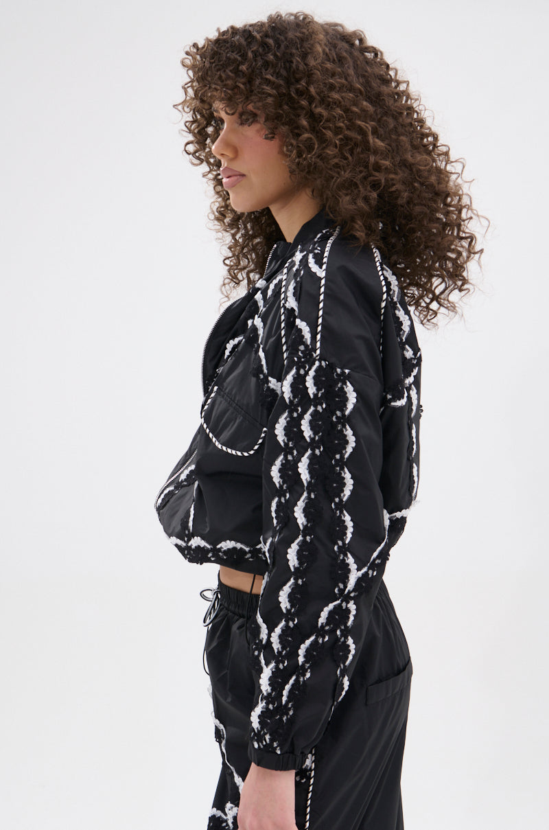 A woman with curly hair models the STRIKE OUT LACE TRIM BOMBER and matching pants, both black with a white wavy line pattern. She stands sideways against a plain white background in a street-casual look.