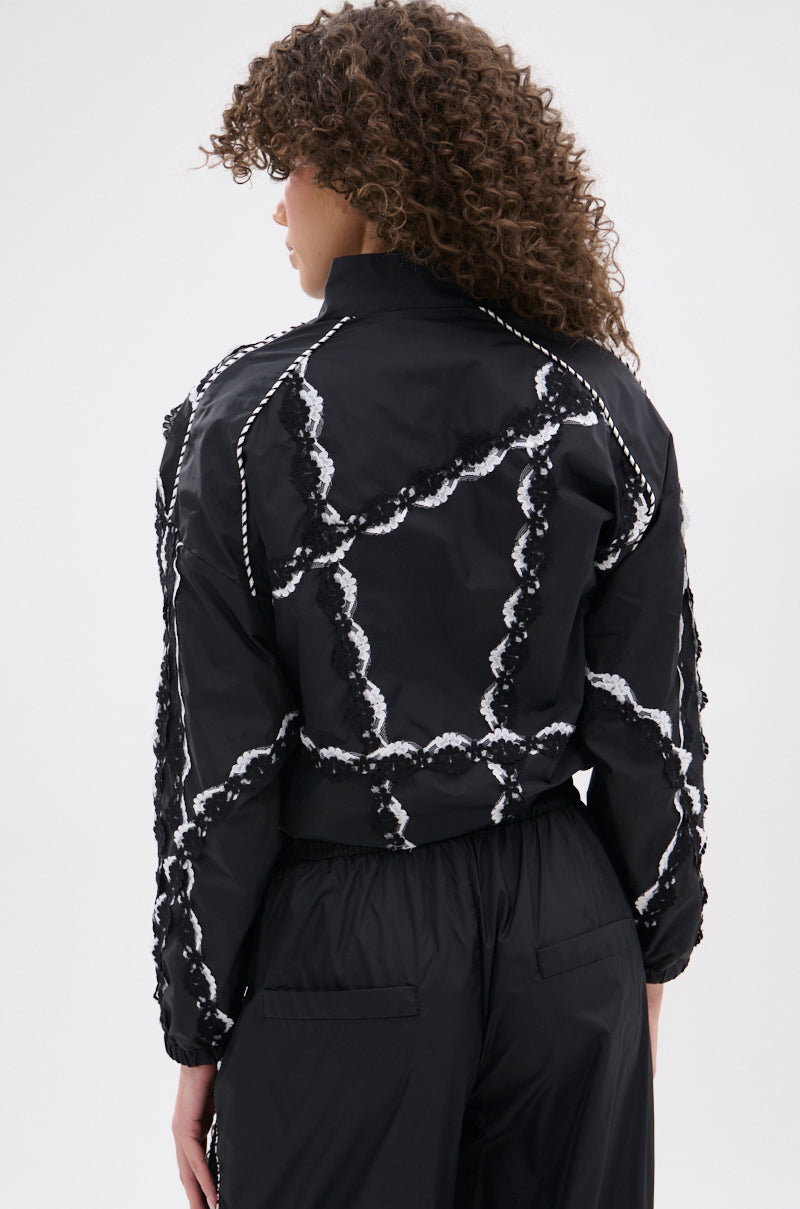 A person with curly hair, seen from behind, wears the STRIKE OUT LACE TRIM BOMBER and wavy-patterned pants in a street-casual style against a plain light background.