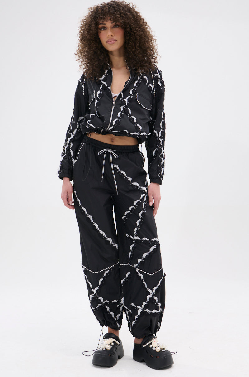 A woman with curly hair models the STRIKE OUT LACE TRIM BOMBER, a street-casual black jacket and pants set with white abstract patterns and black chunky shoes, standing against a plain white background.