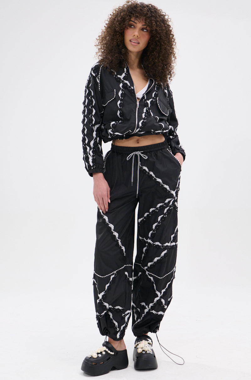A woman with curly hair wears the STRIKE OUT LACE TRIM BOMBER and matching joggers with a white chain pattern, embracing street-casual style. She stands against a plain white background, sporting chunky black shoes and one hand in her pocket.