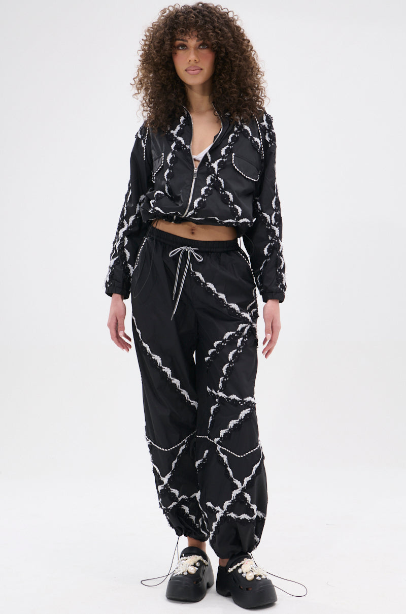 A woman with curly hair models street-casual style in the STRIKE OUT LACE TRIM BOMBER, paired with black crisscross pants, a white crop top, and black platform shoes, standing against a plain white background.