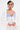 A person with short, slicked-back hair poses confidently against a plain white background, wearing a white outfit with puffy sleeves, a blue-and-white corset top, hoop earrings, visible tattoos, and the COVER UP CHIFFON SHRUG as an open-front layering piece.