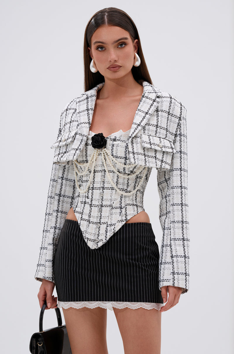 A woman stands against a plain background wearing the HEAVEN SENT CROPPED TWEED BLAZER, styled with a pinstripe mini skirt with lace trim, white hoop earrings, and holding a small black handbag. 1