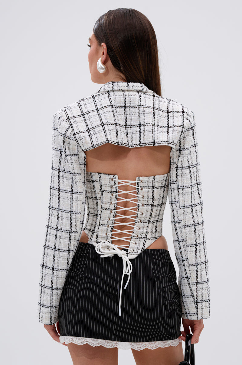 A woman with long brown hair and large white hoop earrings is seen from behind wearing the HEAVEN SENT CROPPED TWEED BLAZER, featuring plaid tweed, pearl buttons, and a white lace-up back, paired with a black pinstripe skirt. 3