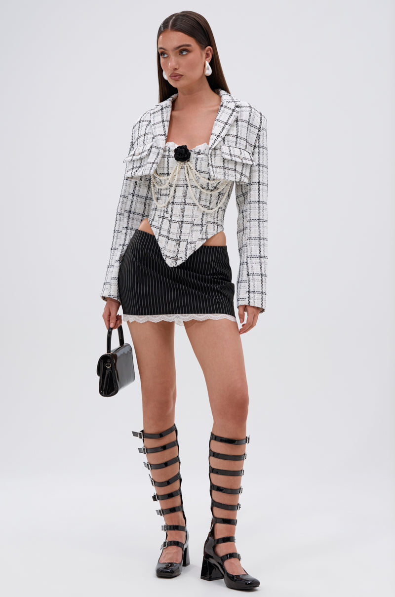 A woman models the HEAVEN SENT CROPPED TWEED BLAZER with pearl buttons and metallic thread, styled with a black pinstripe mini skirt, knee-high strappy heels, and a small black handbag against a plain background. 4