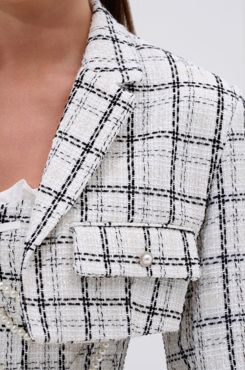 Close-up of the HEAVEN SENT CROPPED TWEED BLAZER in cream and black plaid with pearl buttons, metallic thread detailing, and a flap pocket. A white ruffled top peeks out from beneath the blazer’s upper chest and shoulder. 6