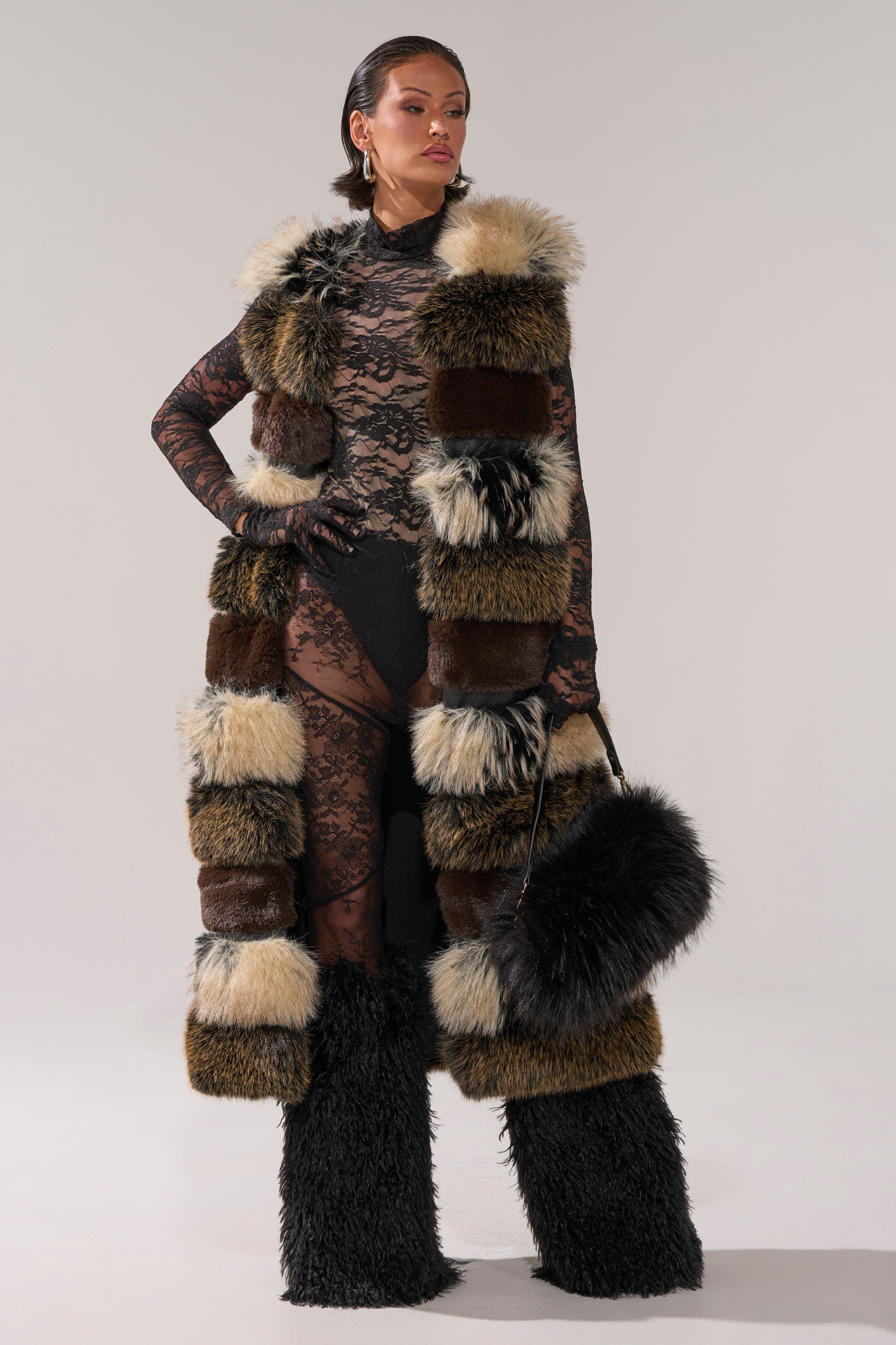 A woman poses in the PORTIA PANELED FUR VEST, featuring a patchwork design, paired with a black lace bodysuit, fur-trimmed boots, and a fluffy black handbag against a plain background.