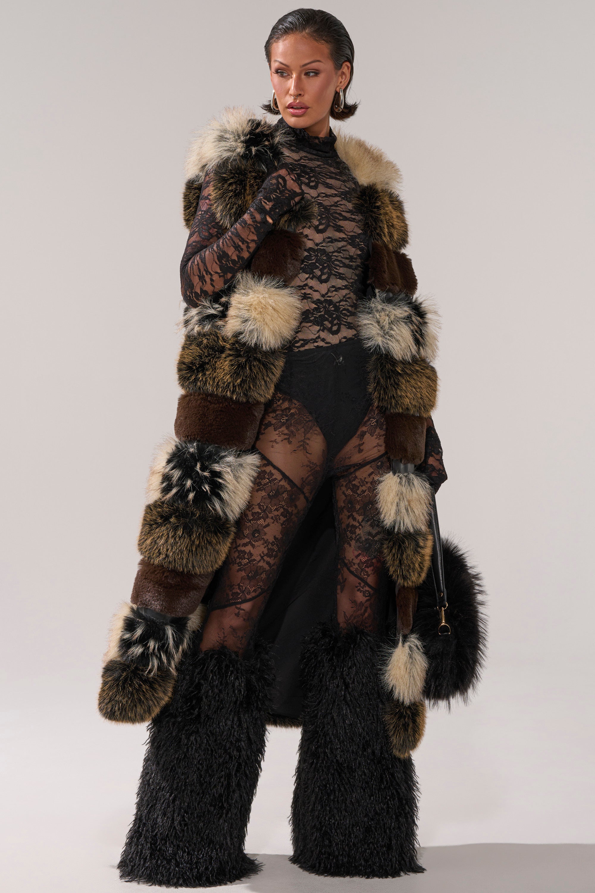 Wearing the PORTIA PANELED FUR VEST over a sheer black lace bodysuit with visible black undergarments, she styles furry black leg warmers, a fur handbag, slicked-back hair, and bold earrings.