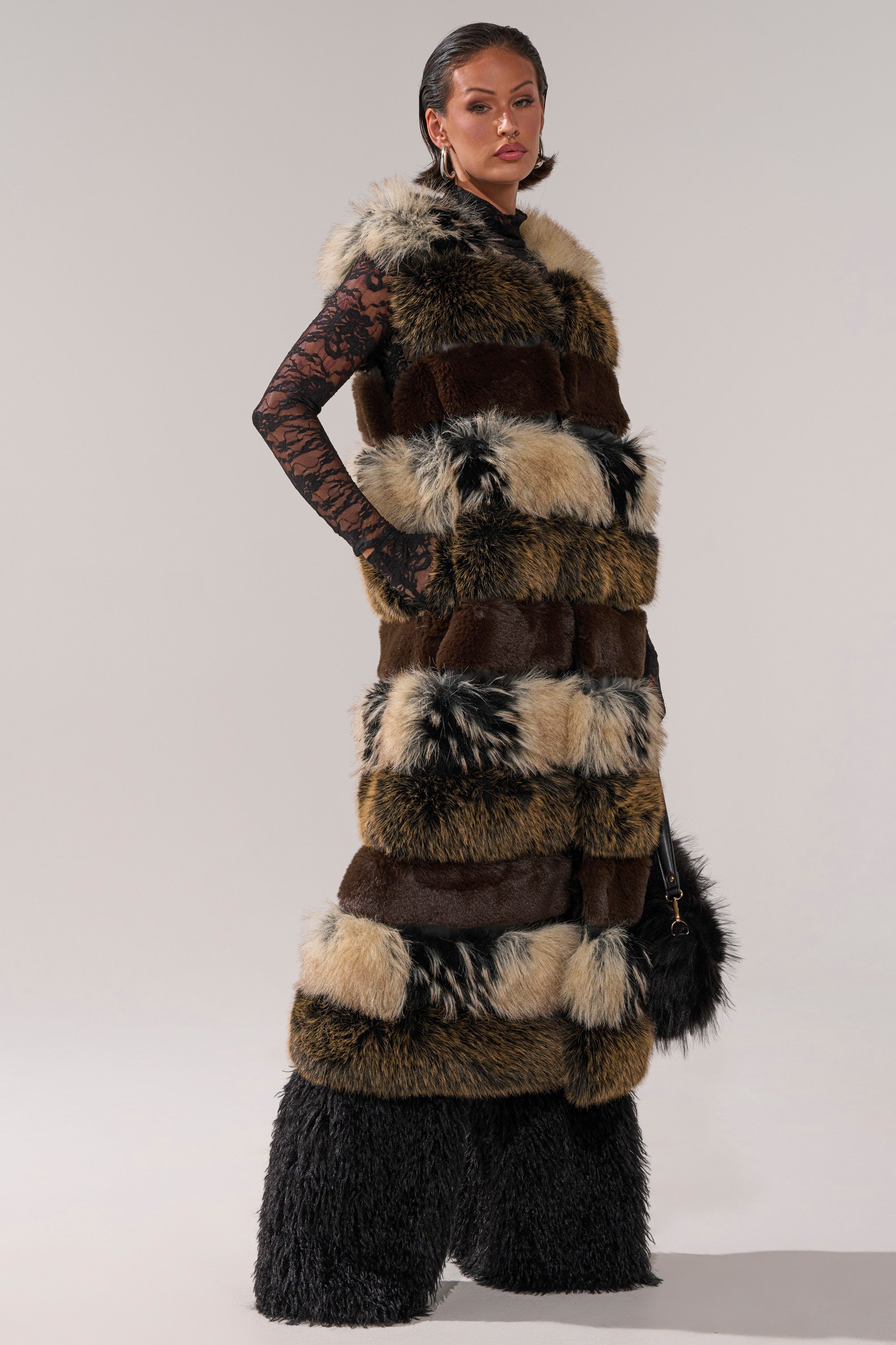 A woman wears the PORTIA PANELED FUR VEST with brown, black, and cream patches over black lace sleeves and voluminous black furry pants. She carries a small black furry bag and has slicked-back hair.