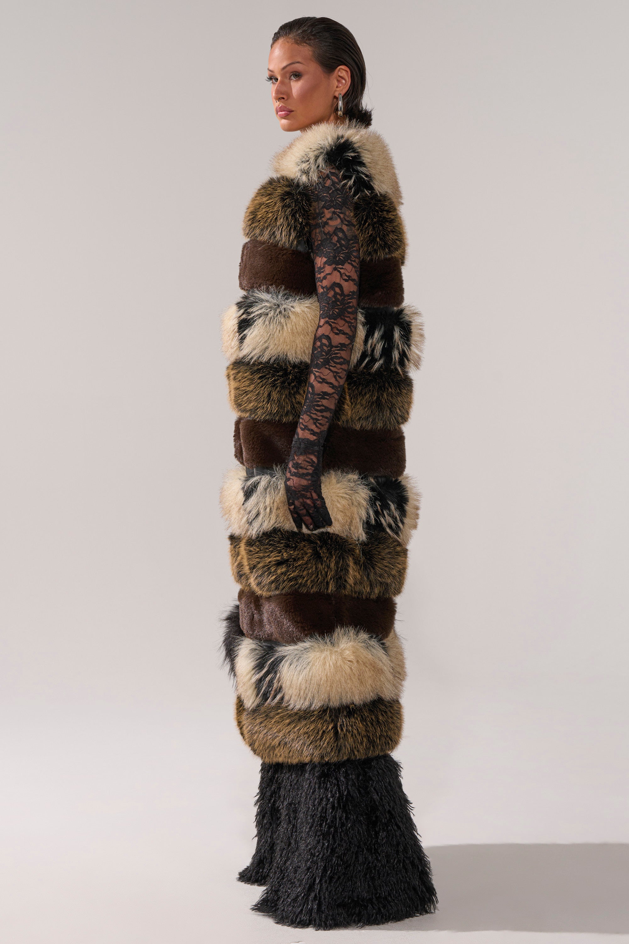 A woman in profile wears the PORTIA PANELED FUR VEST, featuring brown, black, and beige faux fur bands, layered over a black lace-sleeve top and black furry pants. The scene is set against a plain light gray background.