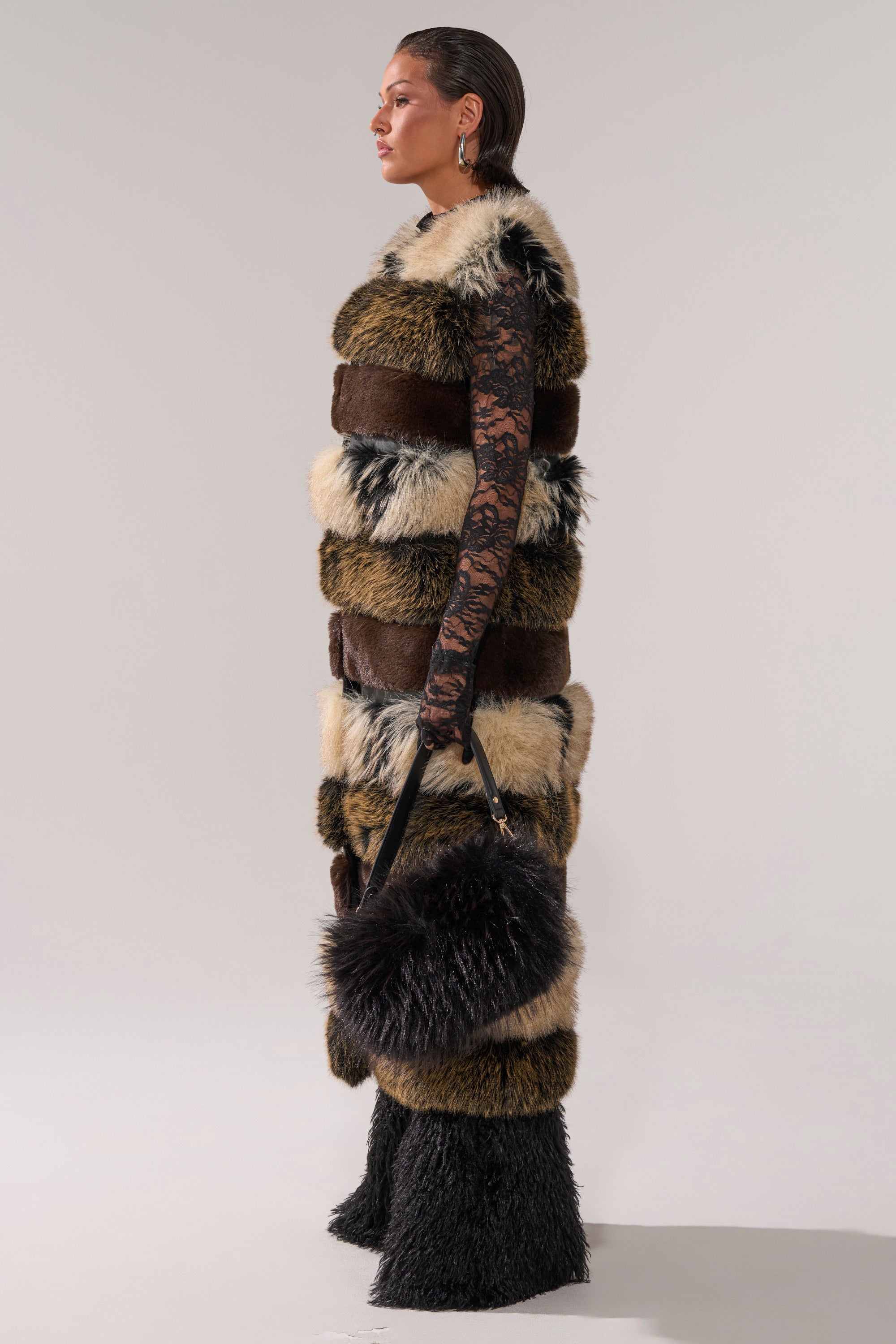A person in profile wears the PORTIA PANELED FUR VEST with brown, black, and beige bands, paired with black lace sleeves, furry pants, and a matching fluffy handbag—creating a bold statement against a plain background.