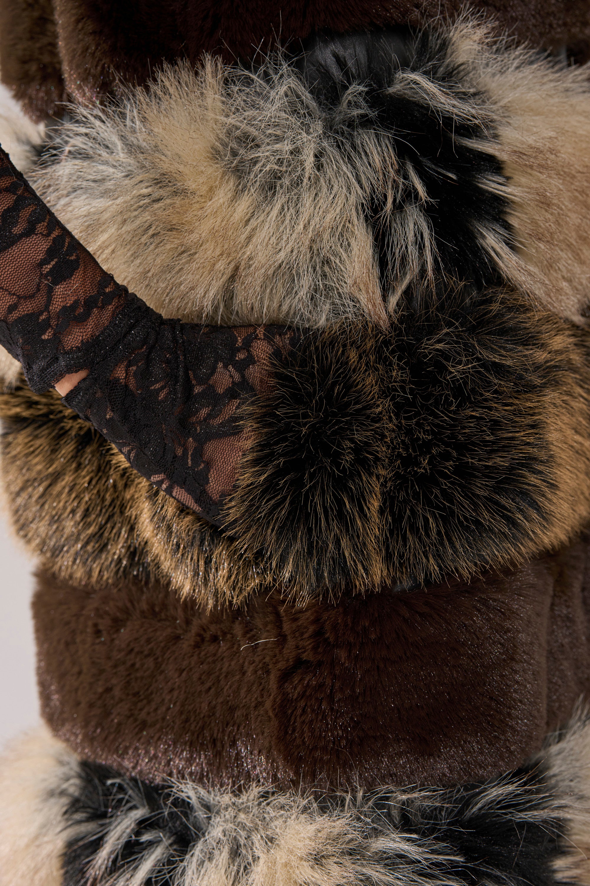 A close-up of a person wearing the PORTIA PANELED FUR VEST, a longline faux fur piece in brown, beige, and black shades, styled with a black lace sleeve crossing over the statement vest.