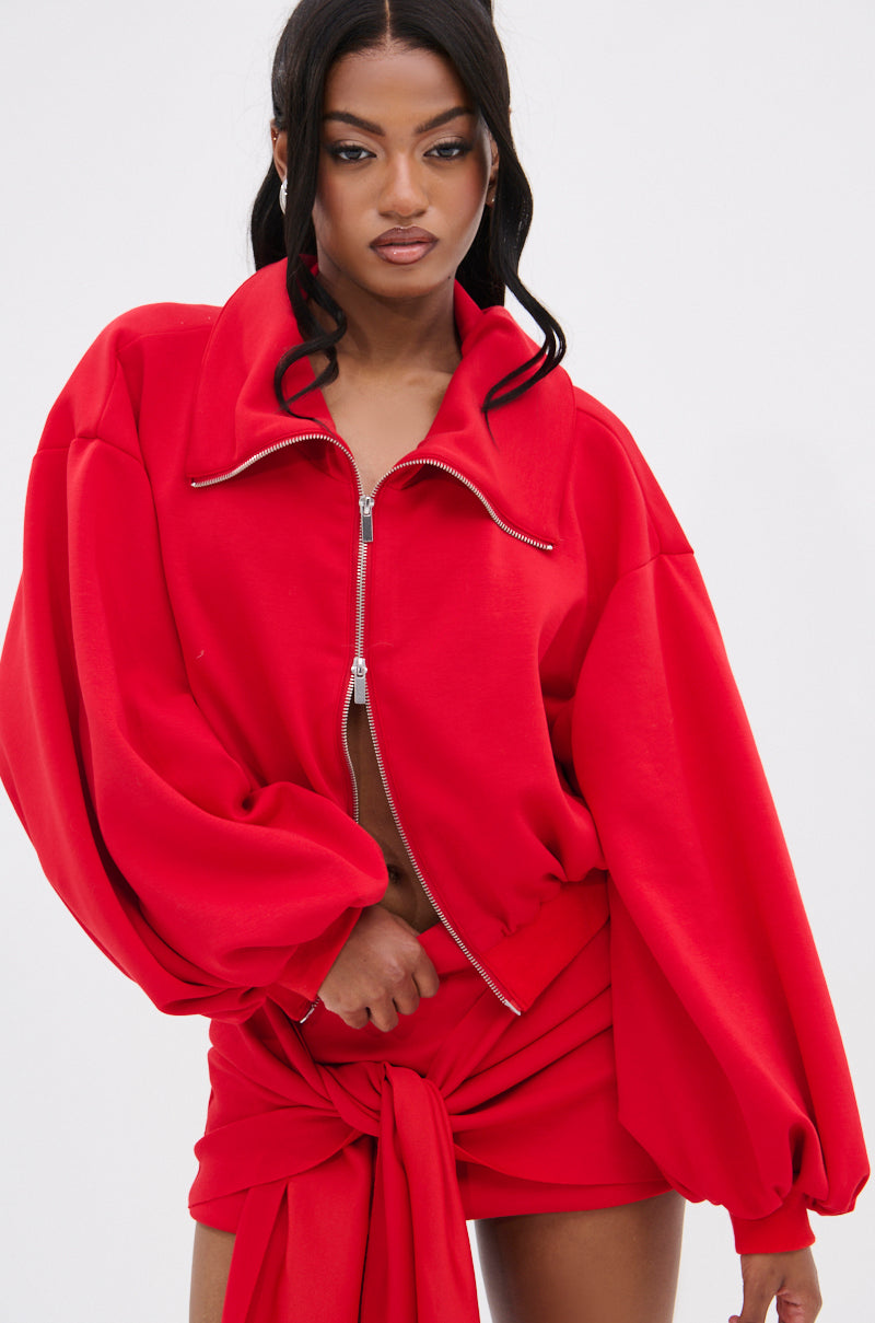 A person with long dark hair poses against a plain background wearing the DON'T BE SHY SCUBA BOMBER in red. The jacket features oversized sleeves and is partially unzipped to reveal their midriff, paired with a matching skirt.