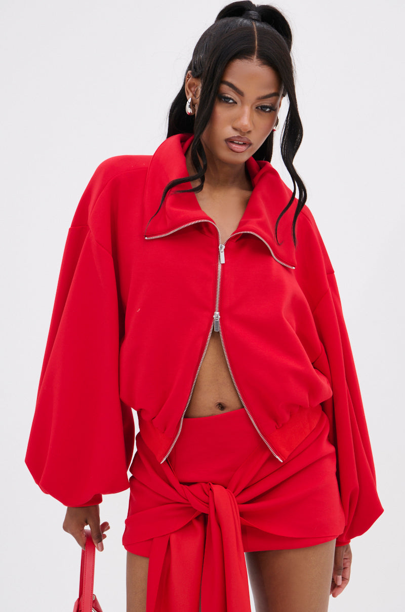 In a striking red AKIRA Label DON'T BE SHY SCUBA BOMBER jacket and skirt with a front knot, a woman carries a matching red bag, her hair styled elegantly in a half-up, half-down ponytail.