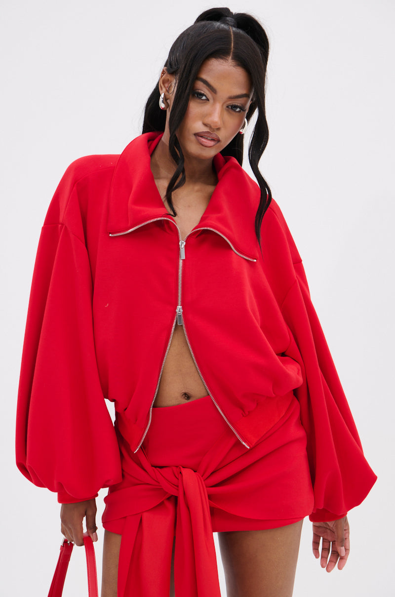 Dressed in the DON'T BE SHY SCUBA BOMBER from AKIRA Label, an individual wears a striking red oversized zip-up top with puff sleeves and a matching skirt with a front tie. Their long, dark hair is styled in high pigtails against a plain white background.
