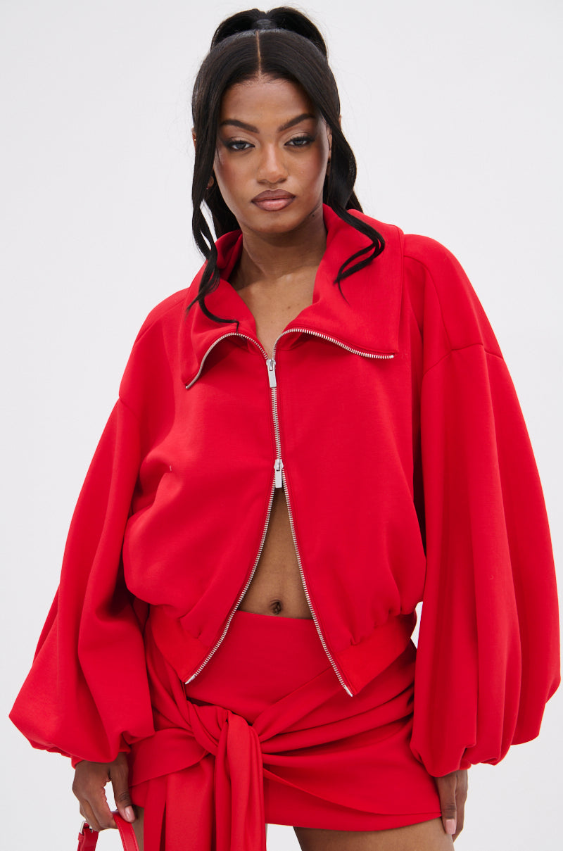 Wearing an AKIRA DON'T BE SHY SCUBA BOMBER jacket with voluminous sleeves, a cropped silhouette, and paired with a wrap-style skirt tied at the waist, the person sports a vibrant red ensemble. Their long dark hair is styled in a high ponytail.