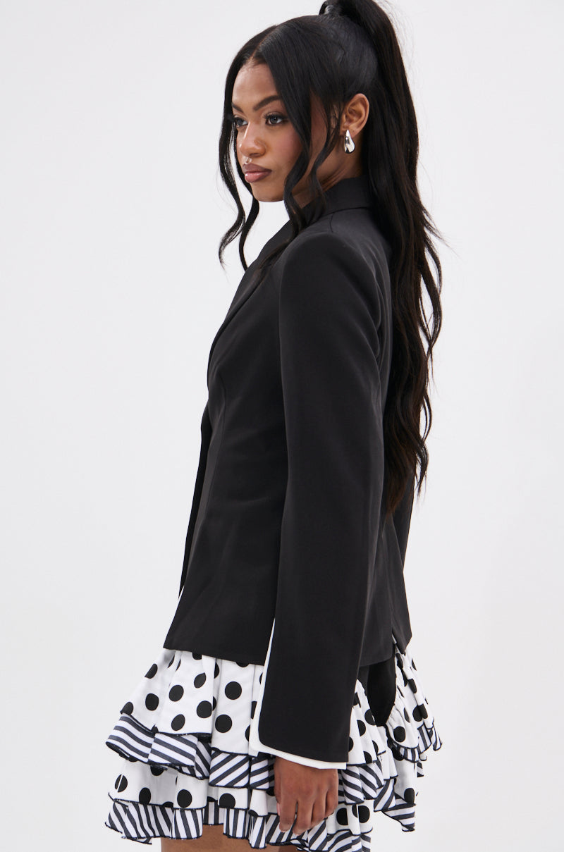 Model in profile wears the TAKE A PEEK DOUBLE LAYER BLAZER over a white skirt with black polka dots and striped ruffles, ideal for transitioning from office to evening, her long wavy black hair stands out against a plain white background.