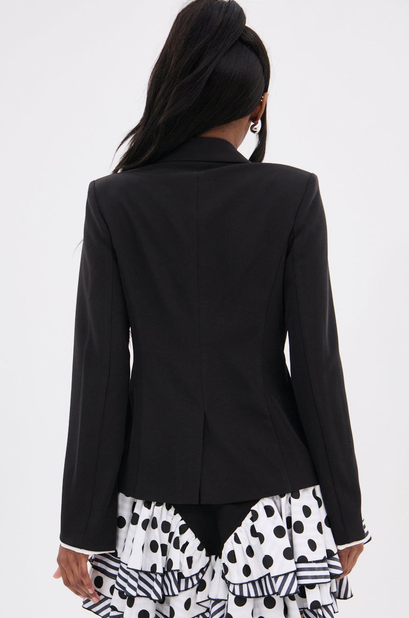A person with long dark hair is shown from the back wearing the TAKE A PEEK DOUBLE LAYER BLAZER over a white polka dot ruffled dress—ideal for transitioning from office to evening. The background is plain white.