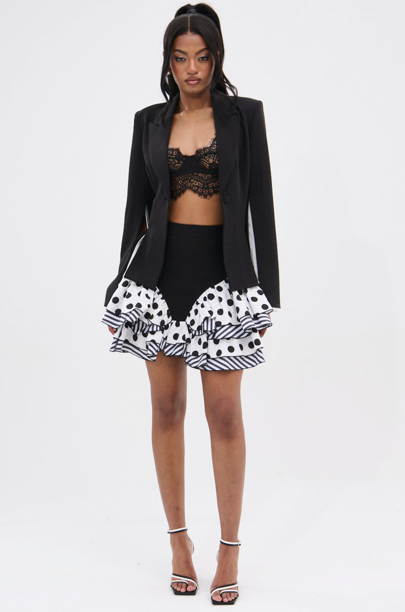 A woman with a high ponytail stands facing forward, wearing the TAKE A PEEK DOUBLE LAYER BLAZER over a black lace bralette, paired with a layered black-and-white polka dot mini skirt and strappy heels—ideal for office-to-evening style.