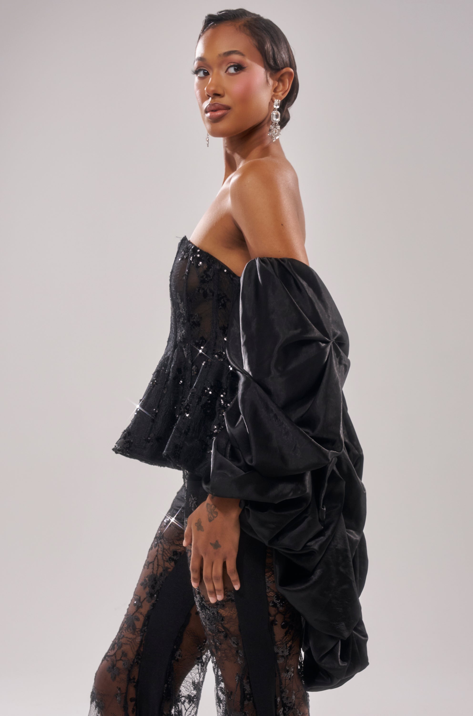 A woman poses against a light gray backdrop, layering the MORE THAN ENOUGH SATIN SHRUG IN BLACK over a chic off-shoulder outfit with sheer lace pants and sparkling earrings. 3