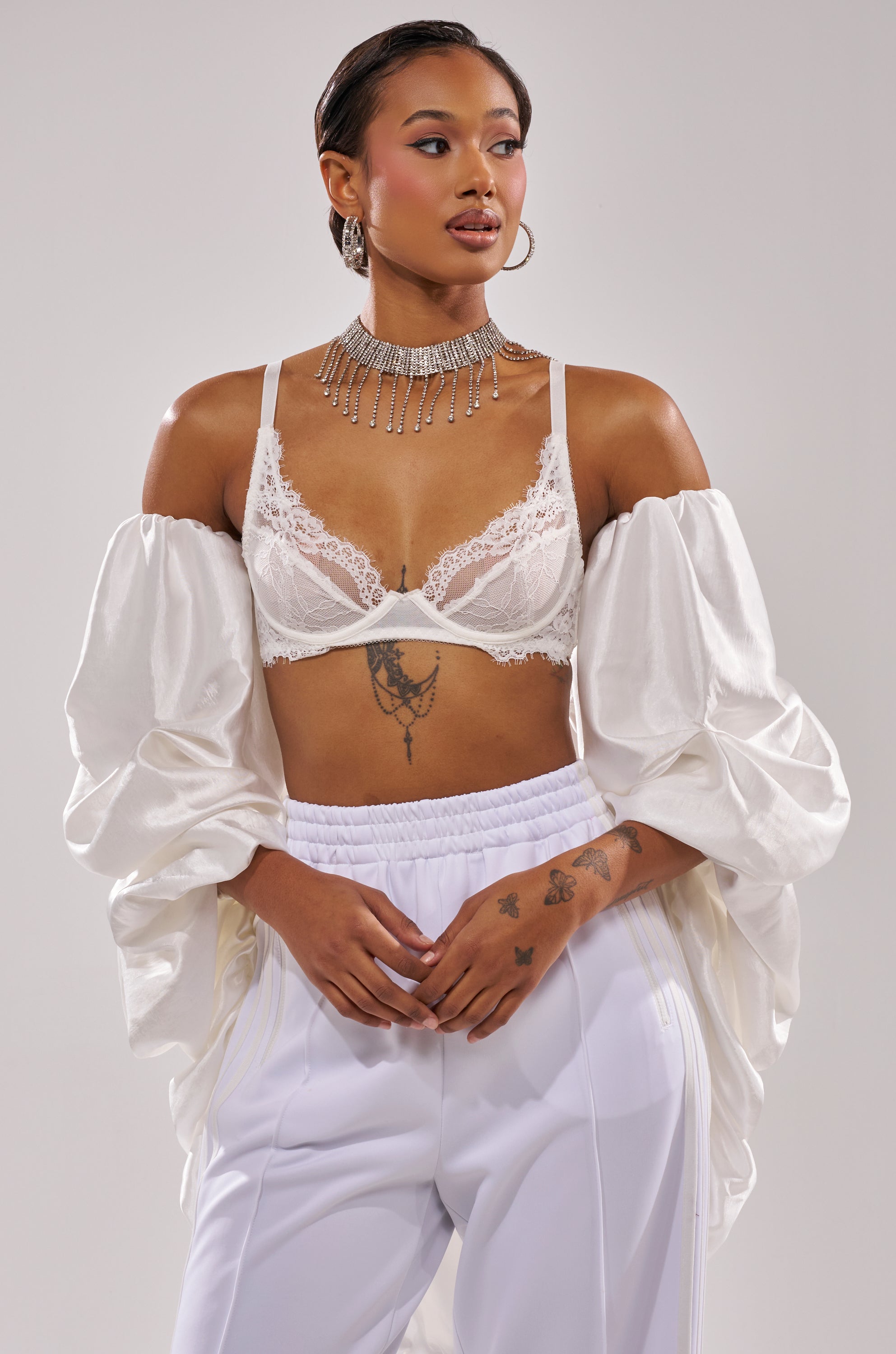 A woman wears the MORE THAN ENOUGH SATIN SHRUG IN WHITE over a lace bralette and white pants, accessorized with a sparkling choker. She stands confidently, hands clasped, displaying her arm and hand tattoos. 1