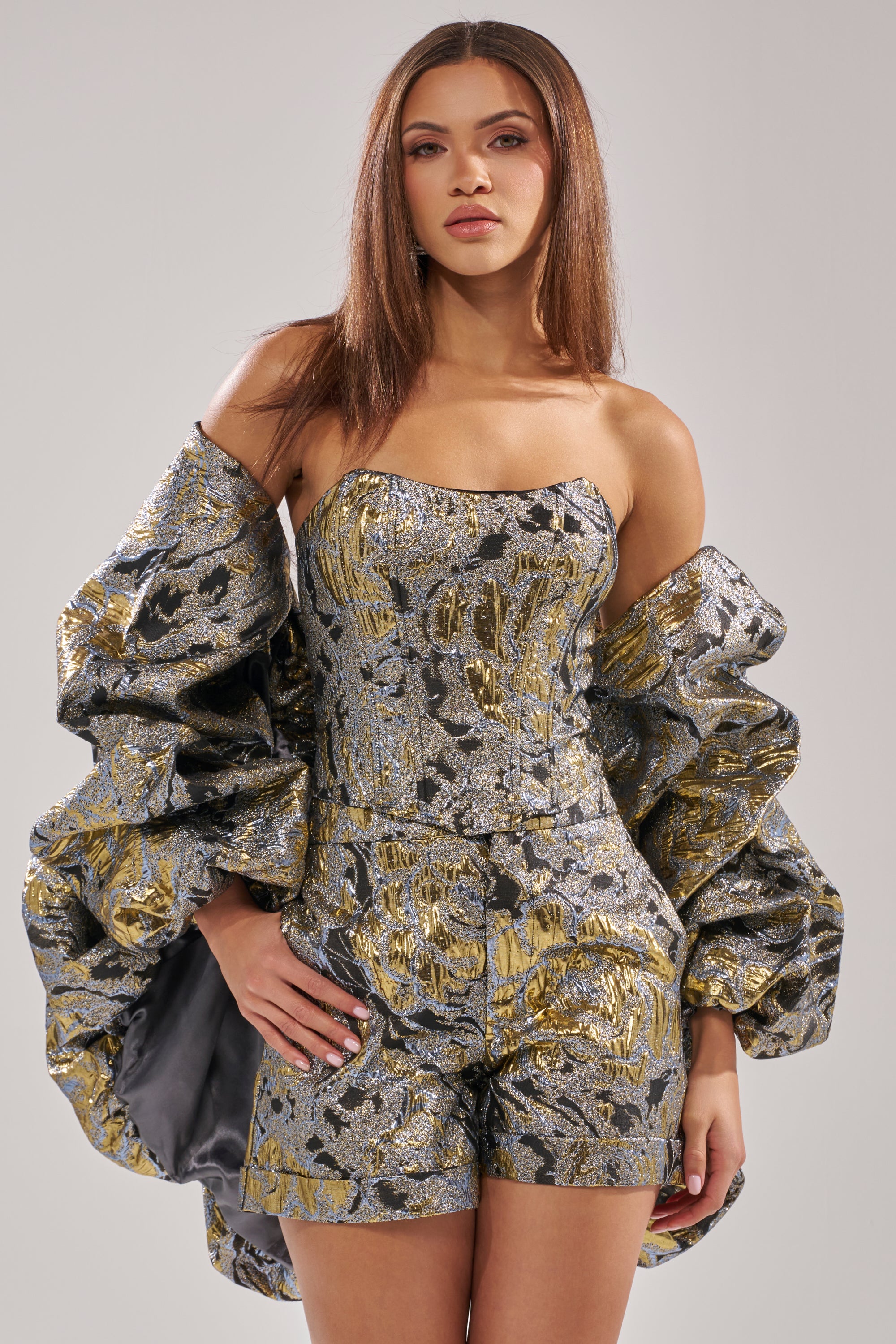 A woman stands confidently against a plain background wearing the AMINA BROCADE SHRUG—metallic silver and gold, with voluminous sleeves—paired with a matching strapless brocade romper.