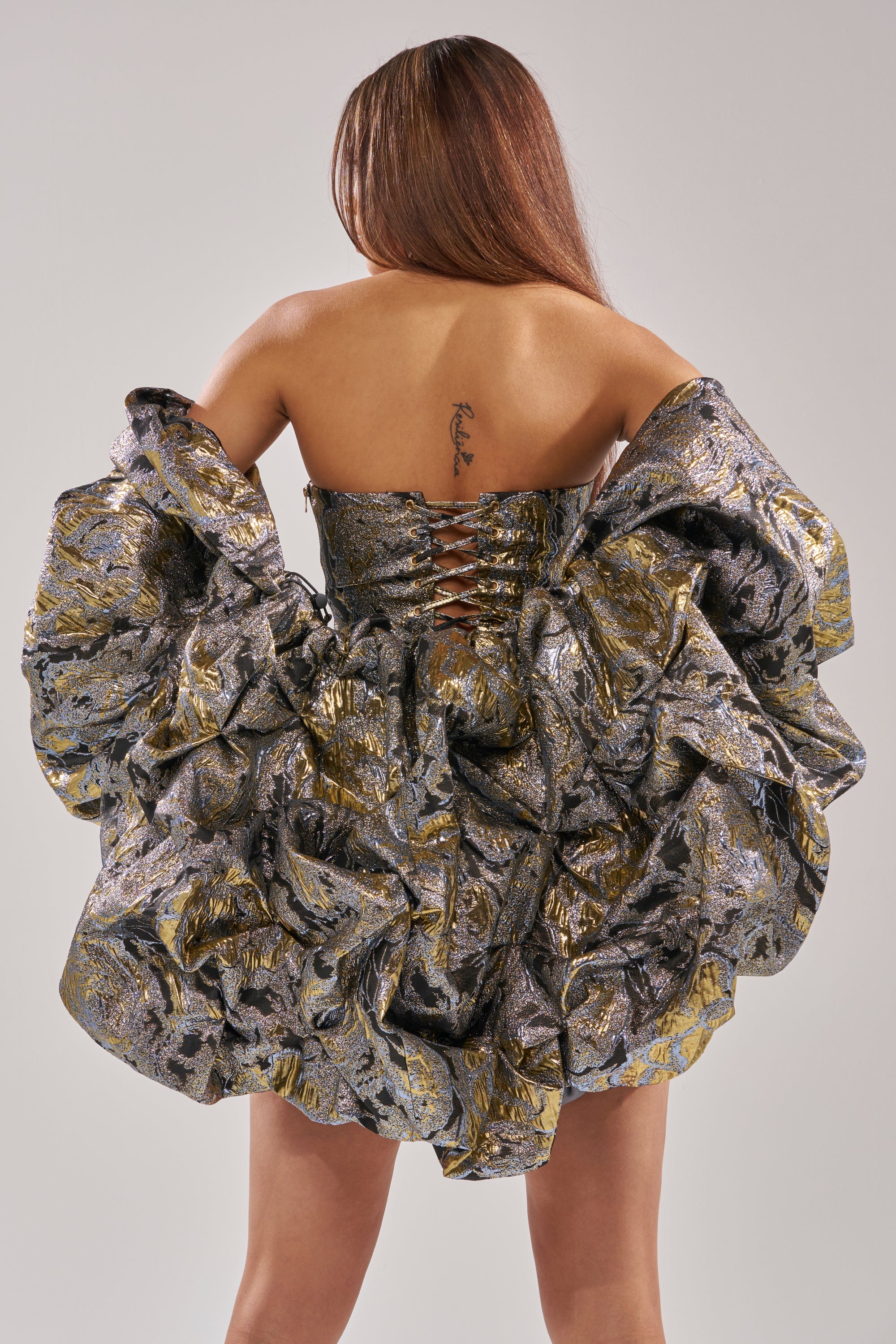 A woman faces away from the camera, wearing the AMINA BROCADE SHRUG over a voluminous metallic silver and gold dress. Her long hair is down, revealing a tattoo and off-the-shoulder sleeves.