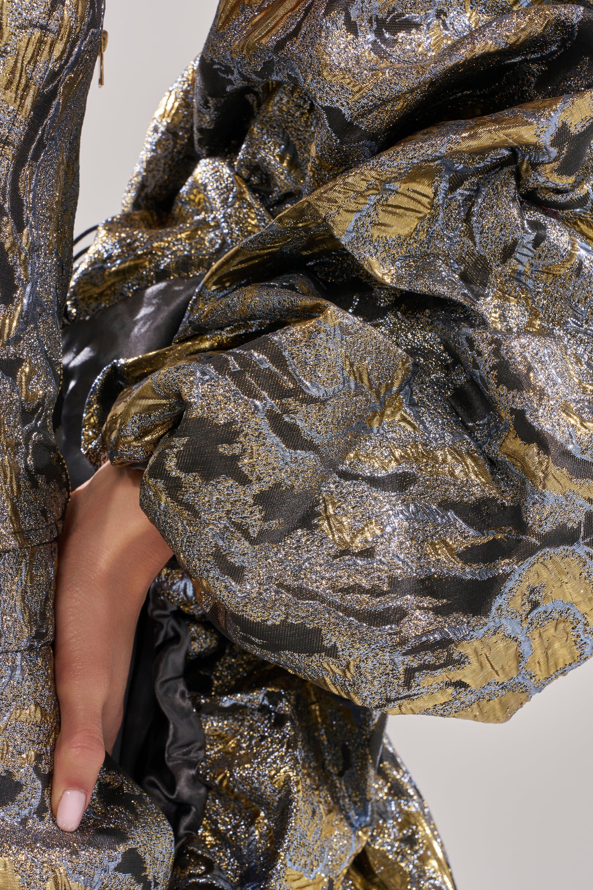 A close-up of a hand resting on the AMINA BROCADE SHRUG, showcasing its metallic gold and silver floral patterns and voluminous, ruffled brocade fabric.