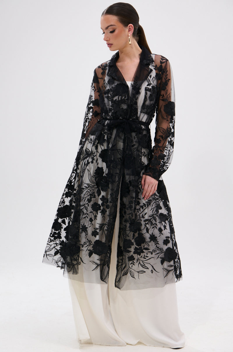 A woman models the IN BLOOM SHEER TRENCH IN BLACK, featuring embroidered floral details, layered over a white floor-length dress against a plain white background for an effortlessly romantic yet rebellious look.
