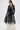 A woman models the IN BLOOM SHEER TRENCH IN BLACK, featuring embroidered floral details, layered over a white floor-length dress against a plain white background for an effortlessly romantic yet rebellious look.