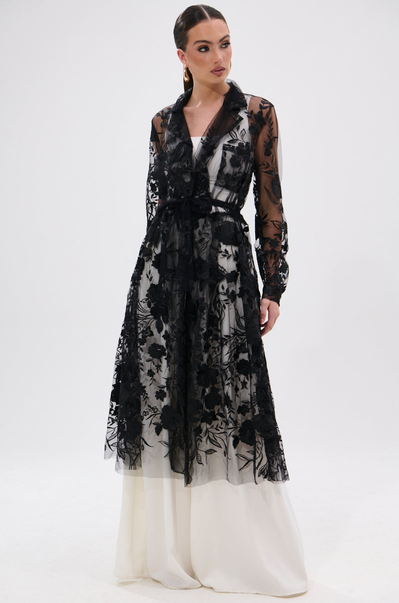 A woman wears the IN BLOOM SHEER TRENCH IN BLACK—a floor-length, embroidered sheer black lace trench with long sleeves over a white layer—standing against a plain white background for a romantic yet edgy look.
