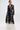 A woman wears the IN BLOOM SHEER TRENCH IN BLACK—a floor-length, embroidered sheer black lace trench with long sleeves over a white layer—standing against a plain white background for a romantic yet edgy look.