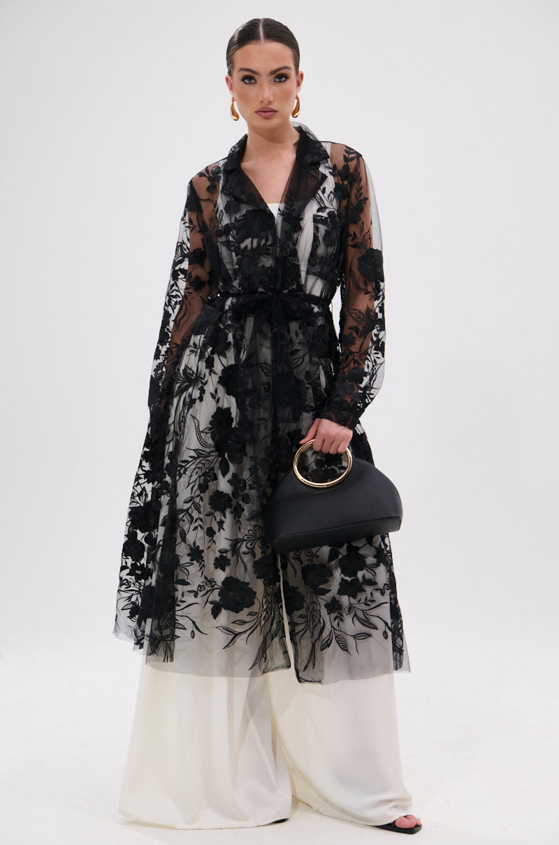 A woman stands against a white background in a romantic, rebellious look, wearing the IN BLOOM SHEER TRENCH IN BLACK with embroidered floral lace over white wide-legged pants. She accessorizes with a black gold-handle handbag and gold hoop earrings.